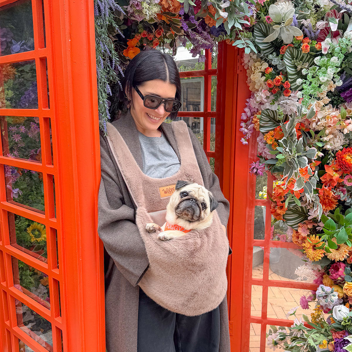 Brown | Furry Range Puppy Pouch: A woman by a red phone box carrying a pug in the Brown Puppy Pouch